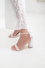 White heeled sandals with a pearl-embellished ankle strap, shown on a model wearing white lace pants against a bright white background.